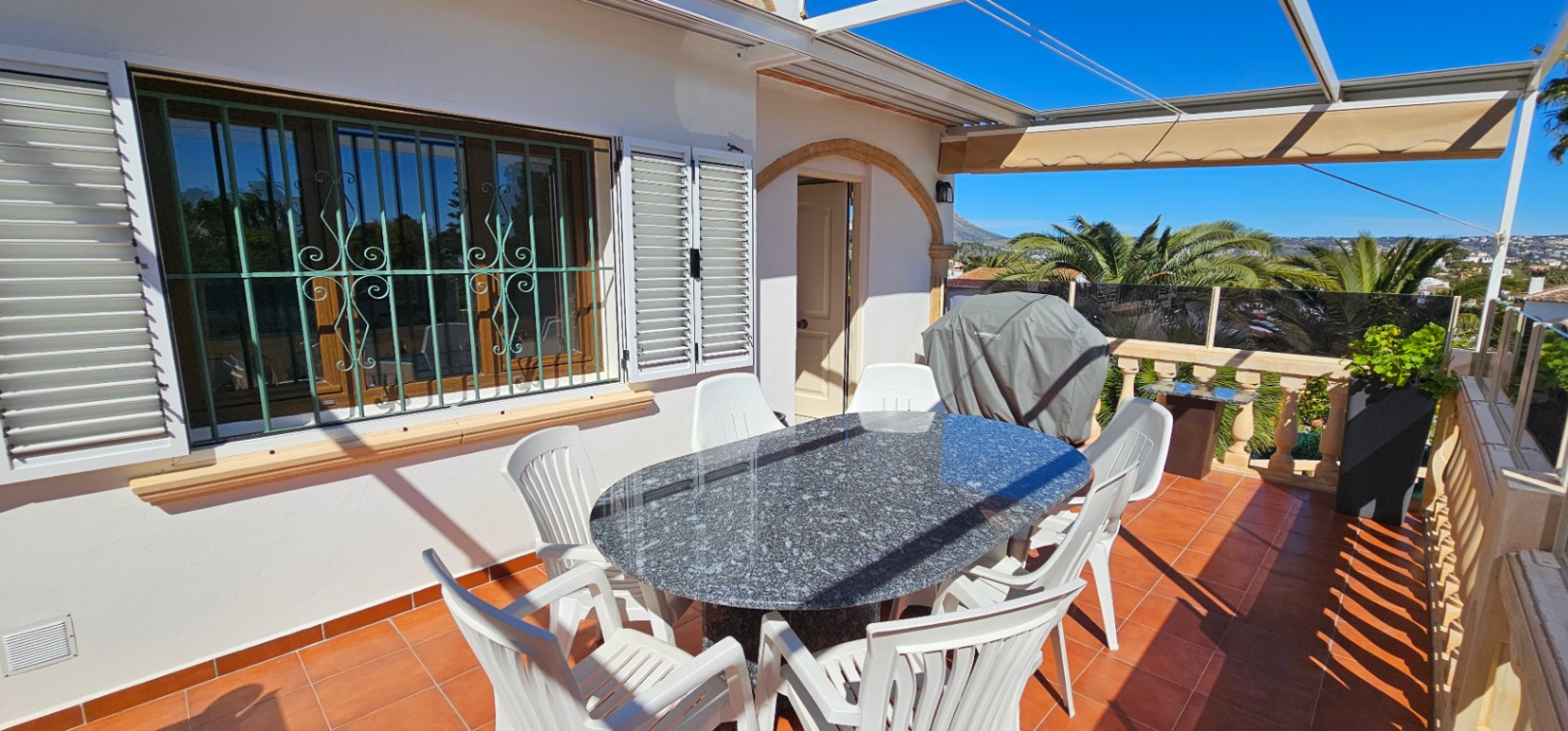 Outdoor terrace featuring terracotta flooring, a granite dining table, and a retractable awning. It includes a stone balustrade and windows with decorative ironwork.
