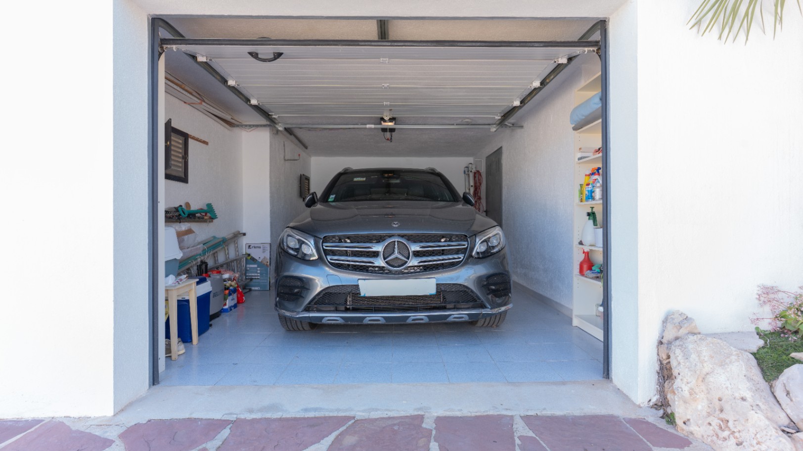 Private garage featuring tiled flooring, integrated shelving, and an automatic sectional door, providing ample storage space and secure vehicle parking.