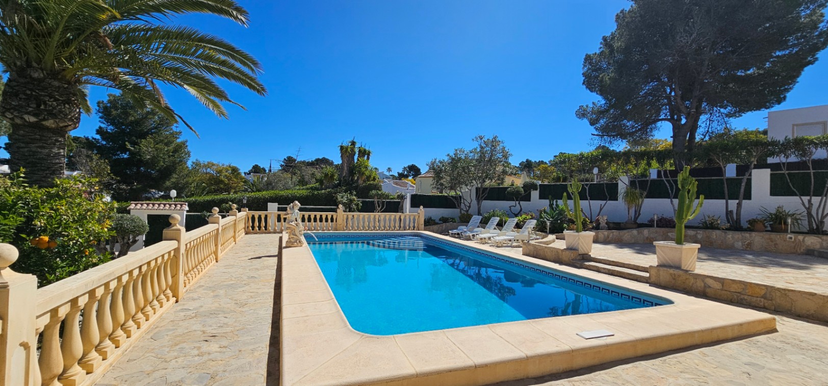 Rectangular swimming pool featuring a stone-paved terrace with a classic balustrade, sun loungers, and lush Mediterranean landscaping under a clear sky.