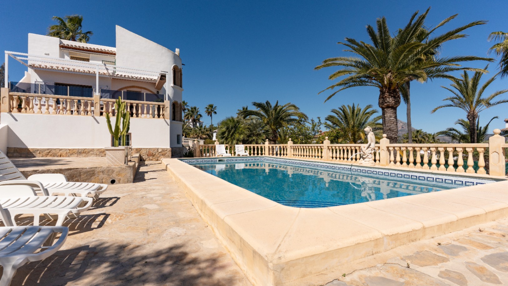 Rectangular swimming pool featuring a stone sun terrace with loungers, surrounded by mature palm trees and the villa's white Mediterranean facade.
