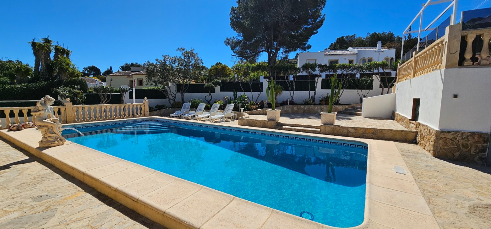 Rectangular swimming pool featuring a stone terrace, white sun loungers, and classical balustrades, framed by Mediterranean gardens and a clear blue sky.