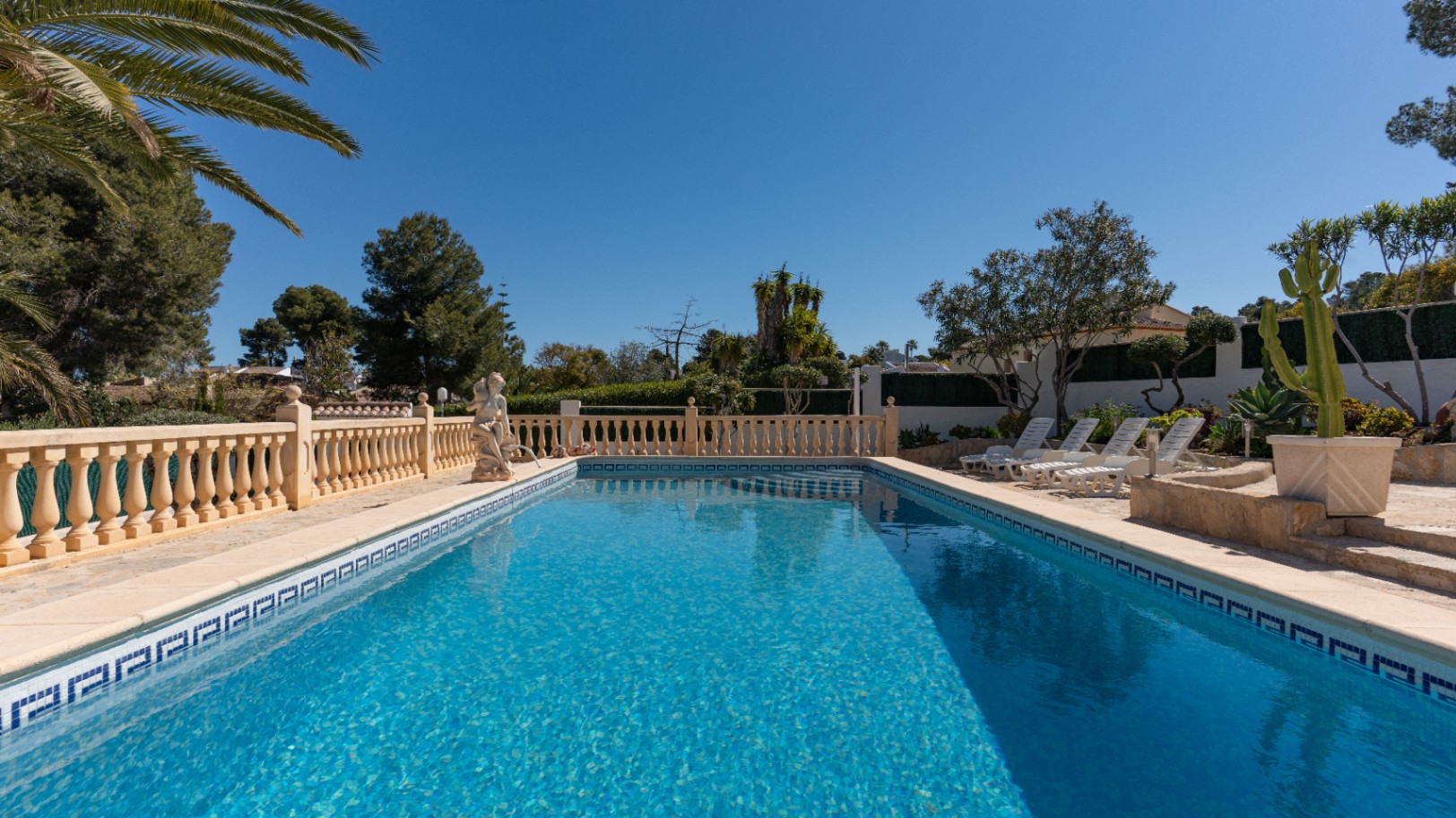 Rectangular swimming pool with blue mosaic tiling, featuring a stone terrace with a classic balustrade, sun loungers, and mature Mediterranean landscaping.