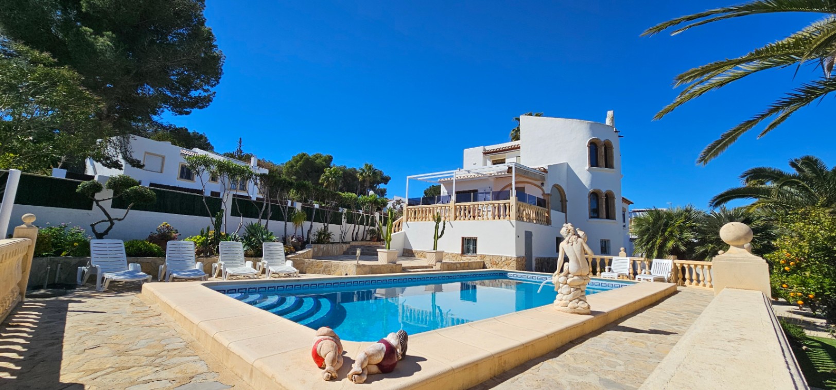 Rectangular swimming pool with Roman steps, surrounded by a stone-paved terrace, white loungers, and a multi-level villa with a rounded tower.