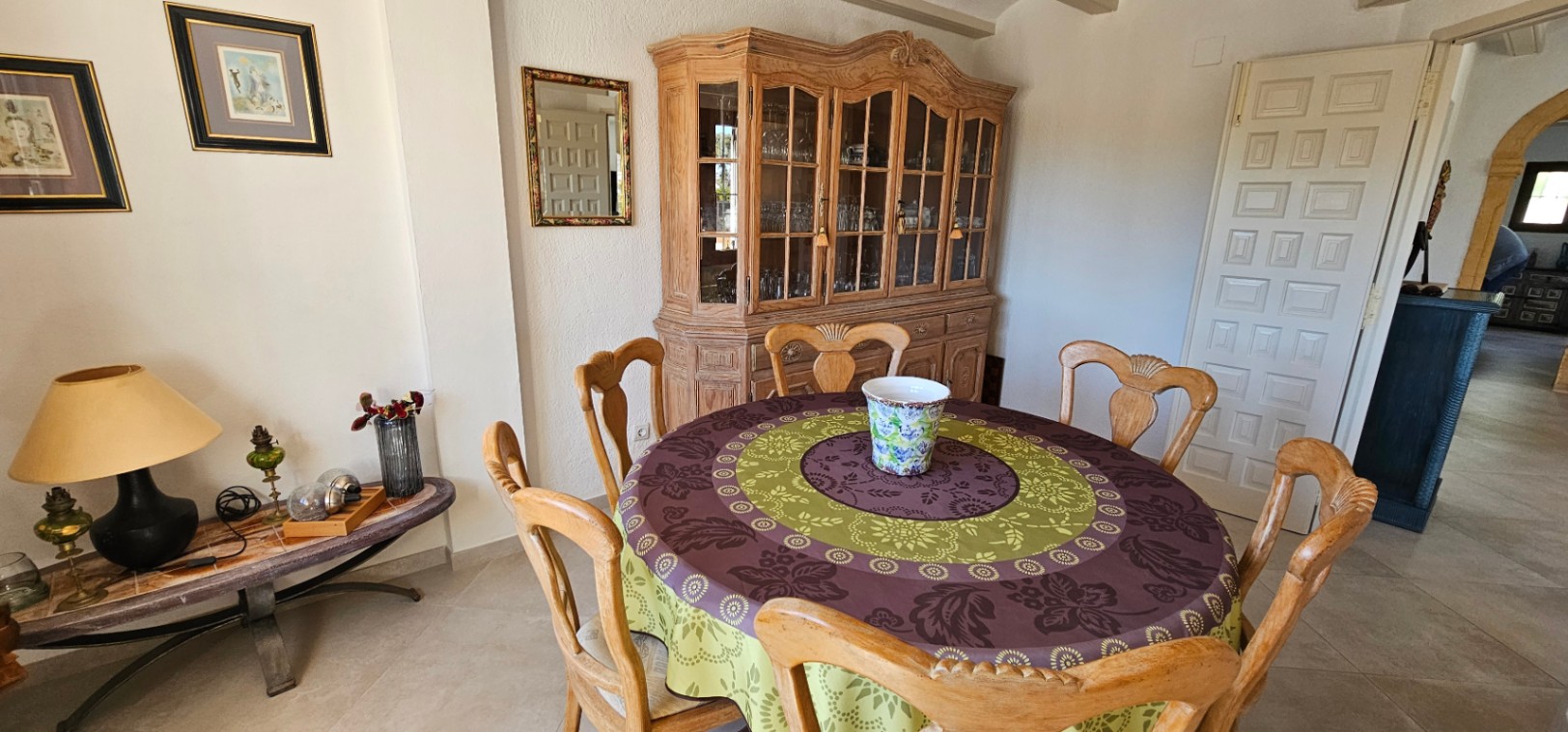 Rustic dining area featuring a round wooden table, carved chairs, and a large traditional display cabinet. The room includes tiled flooring and neutral-toned walls.