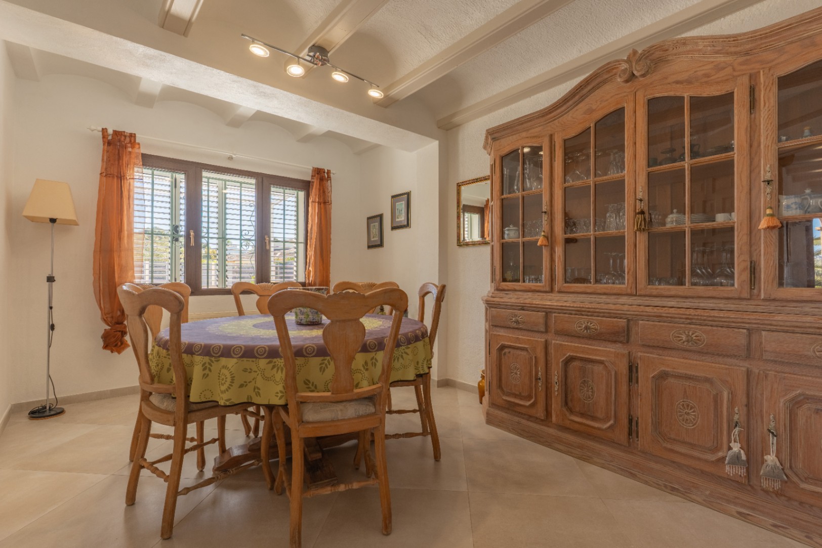 Rustic dining area featuring exposed ceiling beams, a round wooden table, and a large traditional hutch. A shuttered window provides natural light.