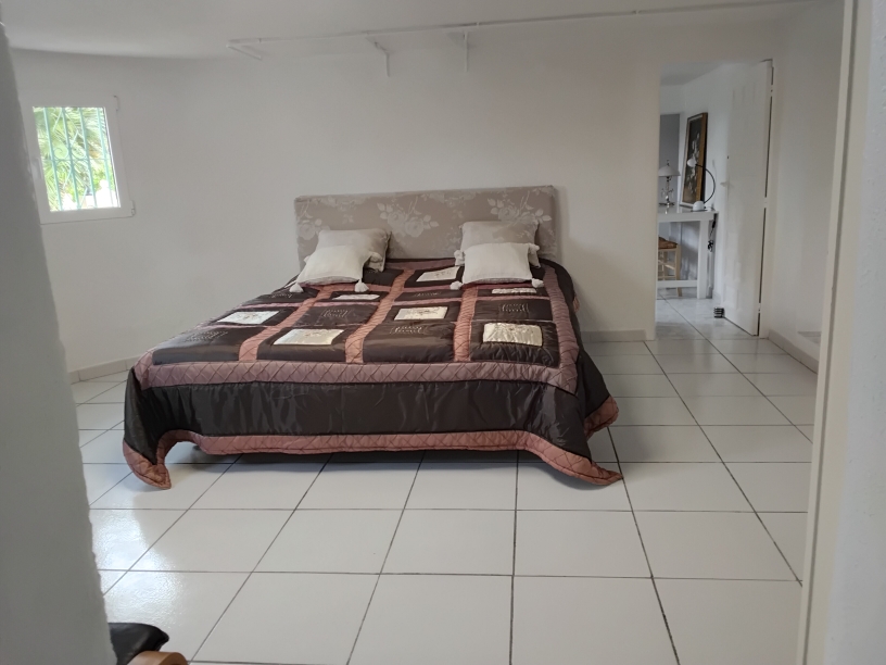Spacious bedroom featuring white tiled flooring, a double bed with a floral headboard, and neutral walls. A small window with security bars provides natural light.