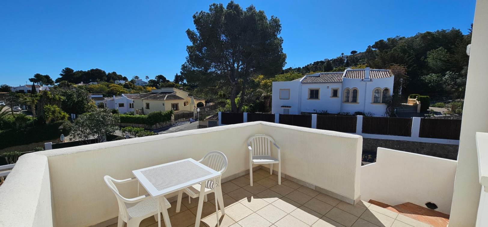 Upper-level open terrace featuring tiled flooring, white outdoor dining furniture, and elevated views of the surrounding residential area and mature trees.