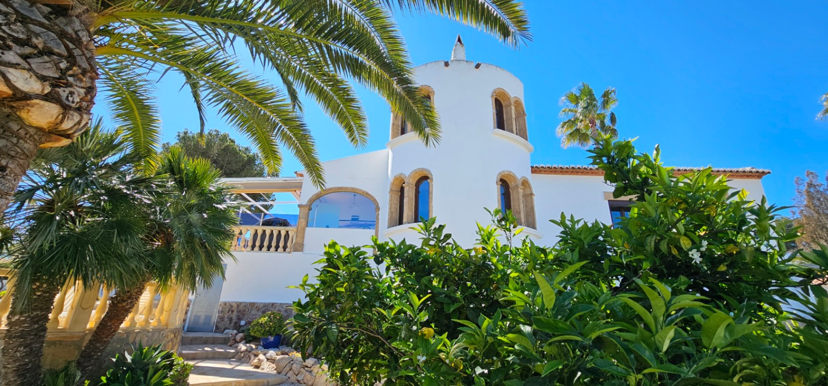 White villa facade featuring a circular tower with traditional Tosca stone arched windows, set within a lush Mediterranean garden with palms and citrus trees.