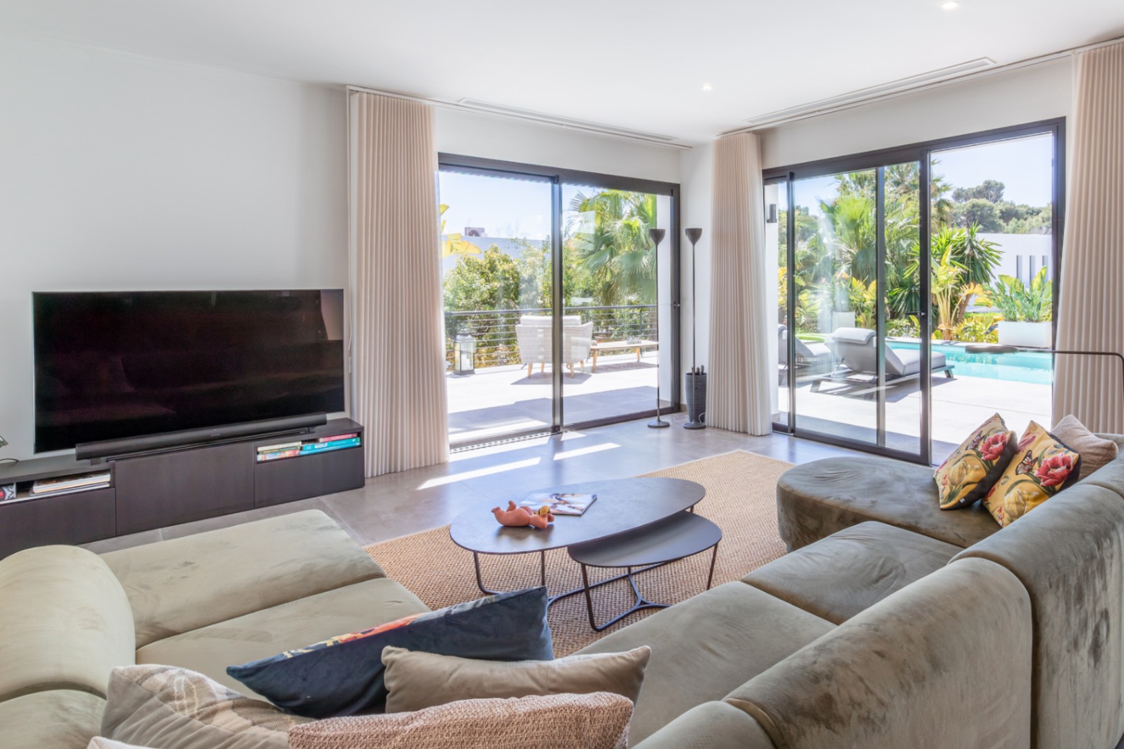 Bright living area featuring a large corner sofa, floor-to-ceiling sliding glass doors opening to the pool terrace, and integrated recessed lighting.