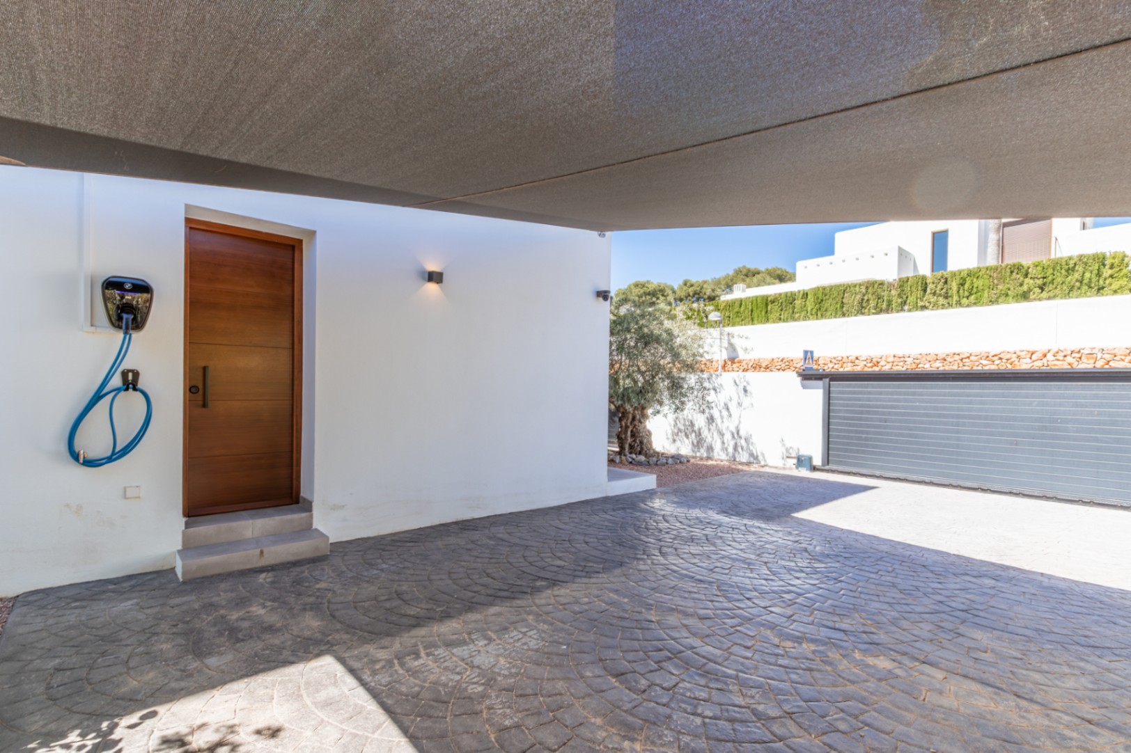 Covered parking area with a shade sail, featuring stamped concrete flooring, a wall-mounted EV charger, and a wooden side door for direct access to the interior.