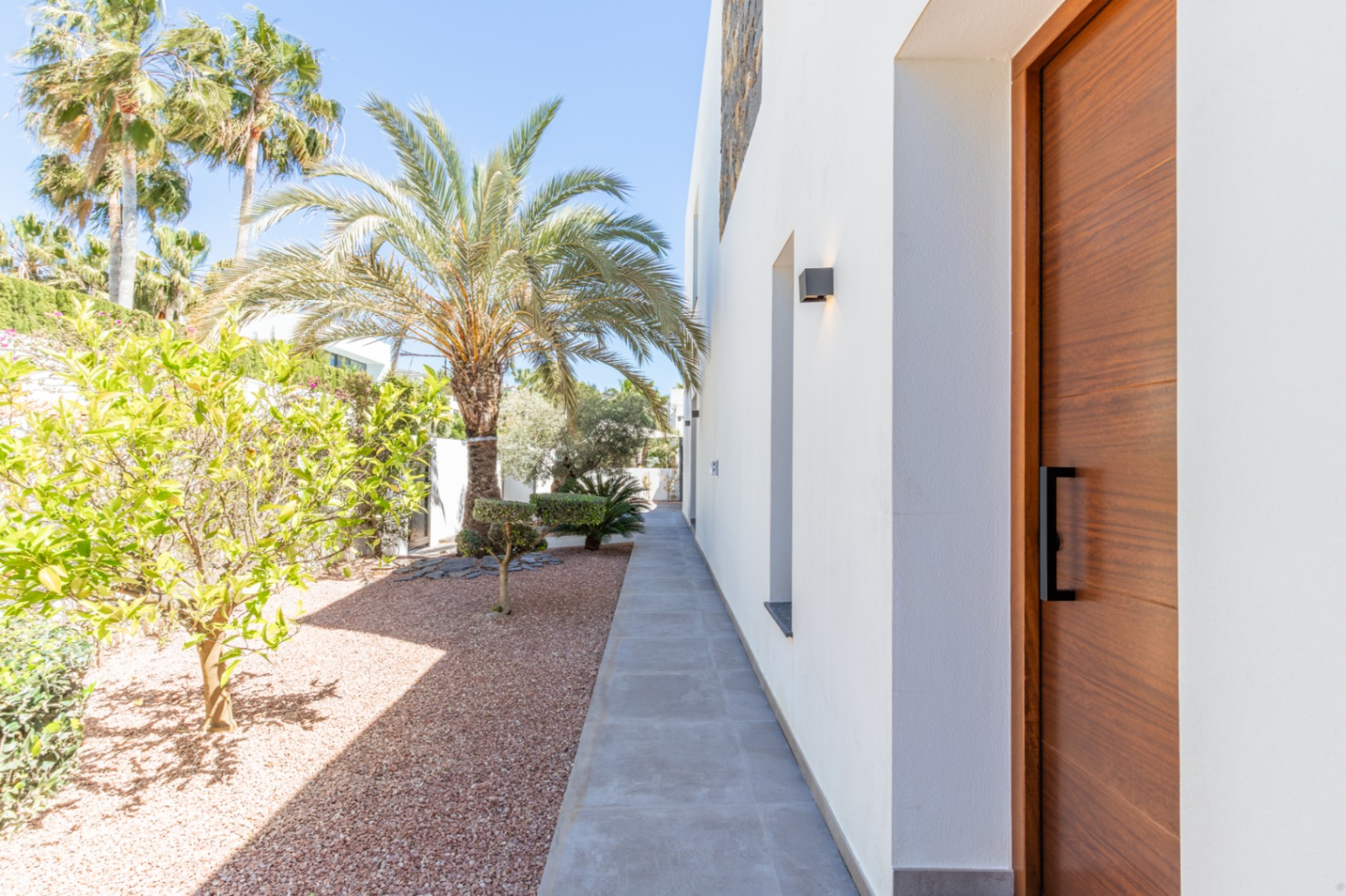 Main entrance featuring a solid wood door and black hardware, adjacent to a paved walkway and a landscaped garden with gravel, palms, and citrus trees.