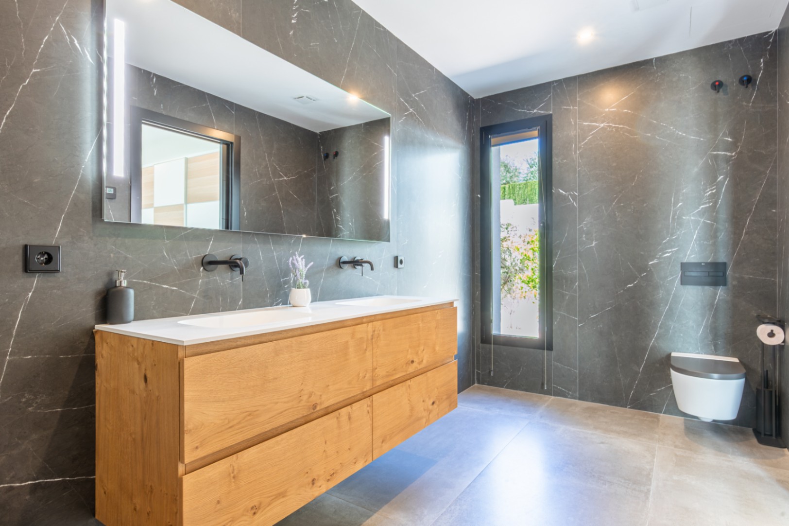Modern bathroom featuring dark marble-effect wall tiles, a double wooden vanity, and matte black faucets. Includes a wall-hung toilet and a large mirror with integrated LED lighting.