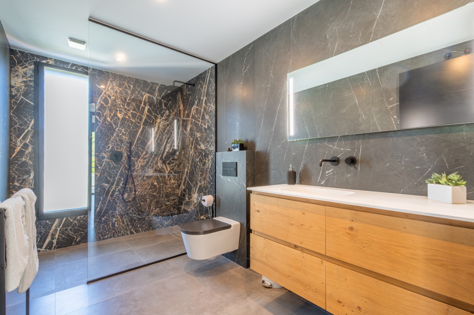 Modern bathroom featuring dark marble-effect wall tiles, a natural wood vanity with white countertop, and a spacious walk-in shower with black fixtures.