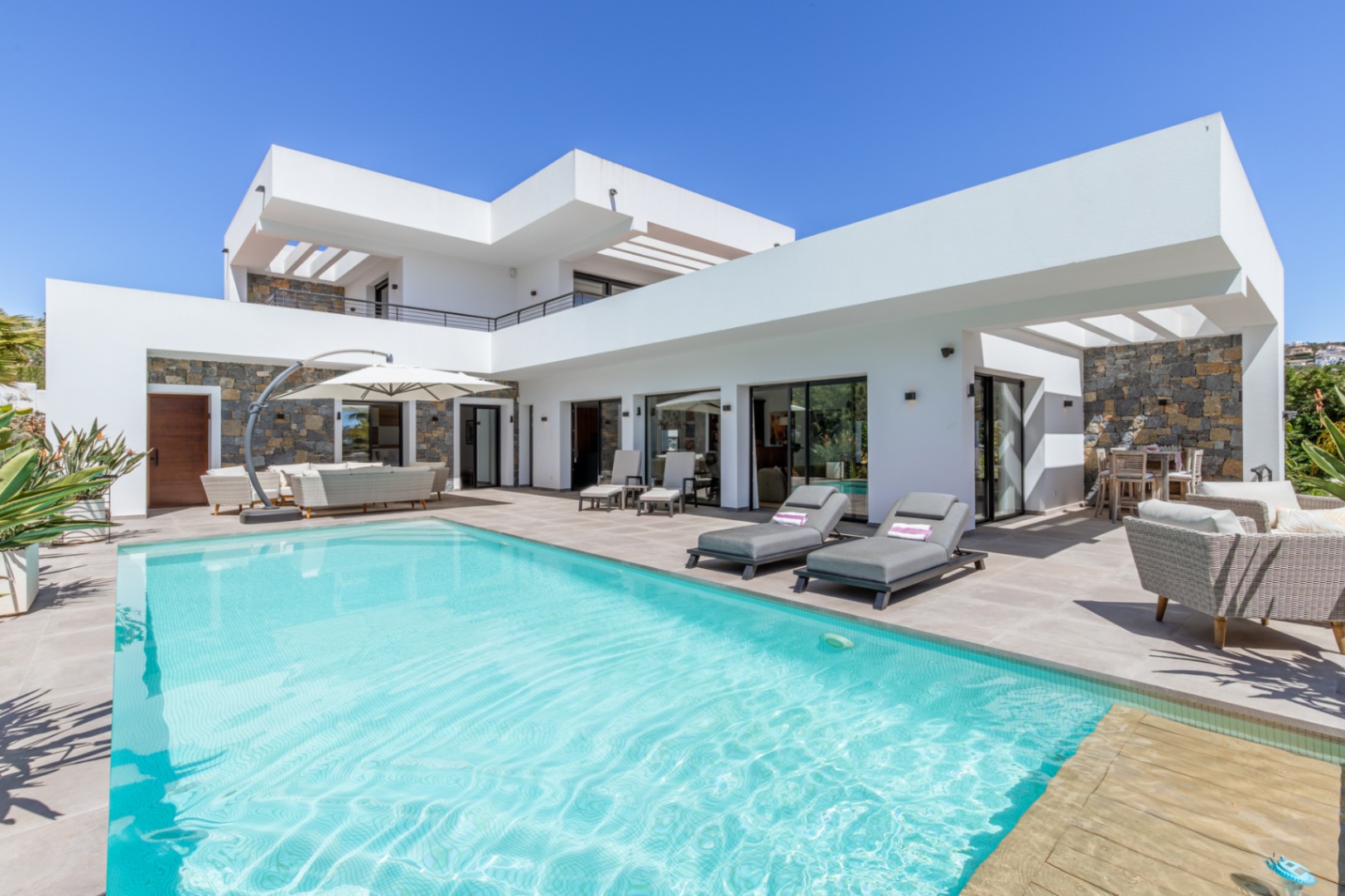 Modern villa exterior featuring a large swimming pool, sun loungers, and a spacious terrace with natural stone wall accents and minimalist architecture.