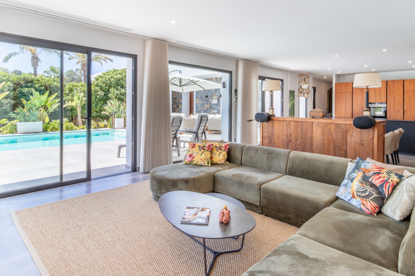 Open-plan living area featuring a green velvet sofa, large sliding doors to the pool terrace, and an integrated kitchen with natural wood cabinetry.