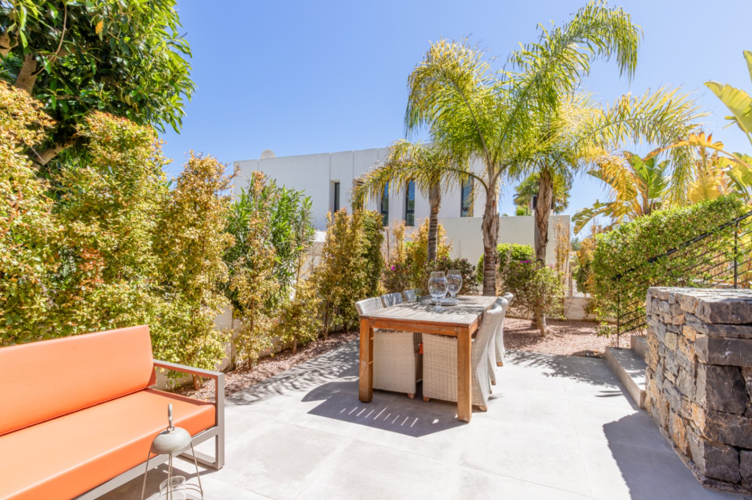Outdoor dining area with a wooden table, wicker chairs, and an orange sofa, set against a backdrop of mature Mediterranean plants and palm trees.