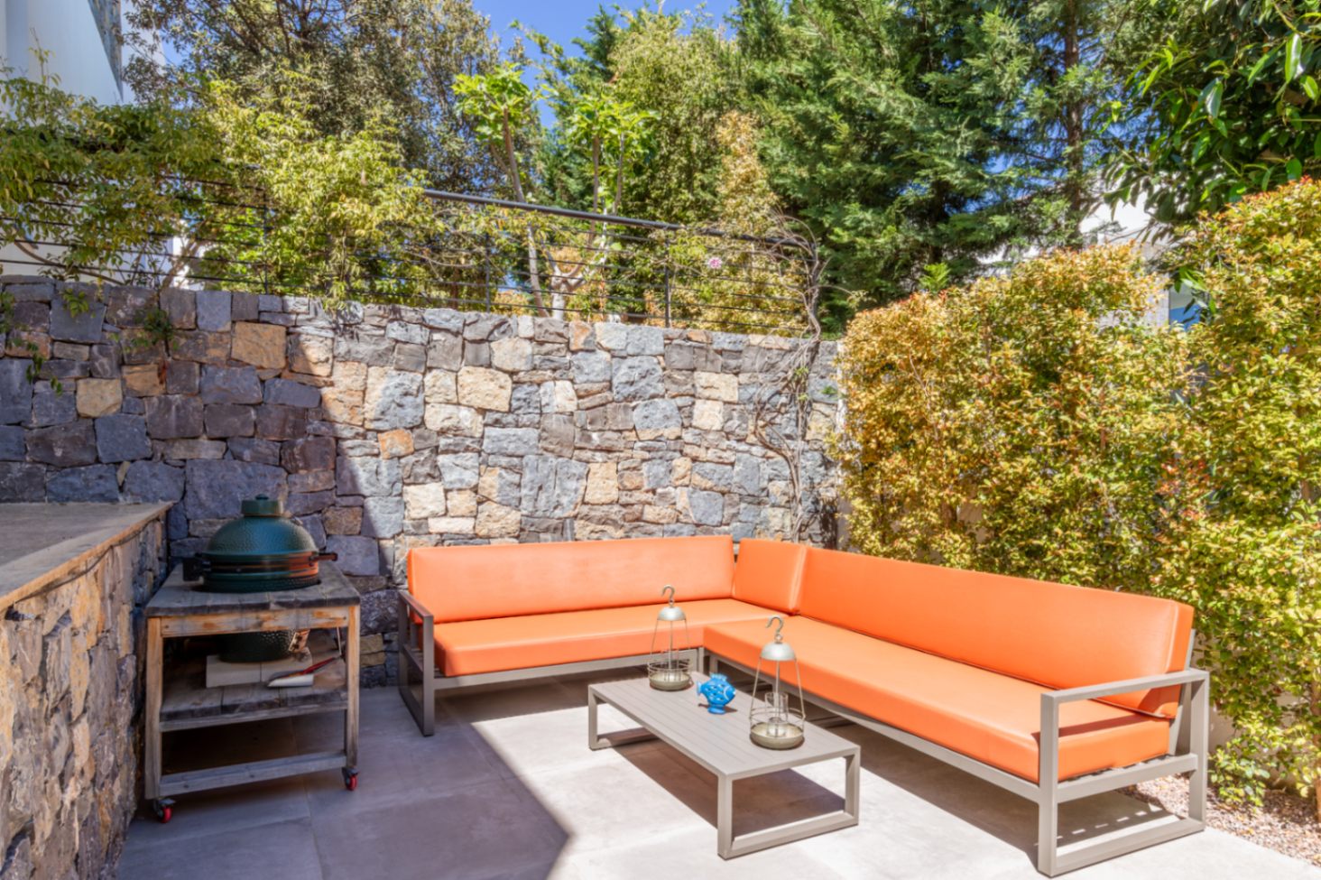 Outdoor lounge area featuring an orange corner sofa, a ceramic kamado grill, and a natural stone wall framed by private Mediterranean greenery.