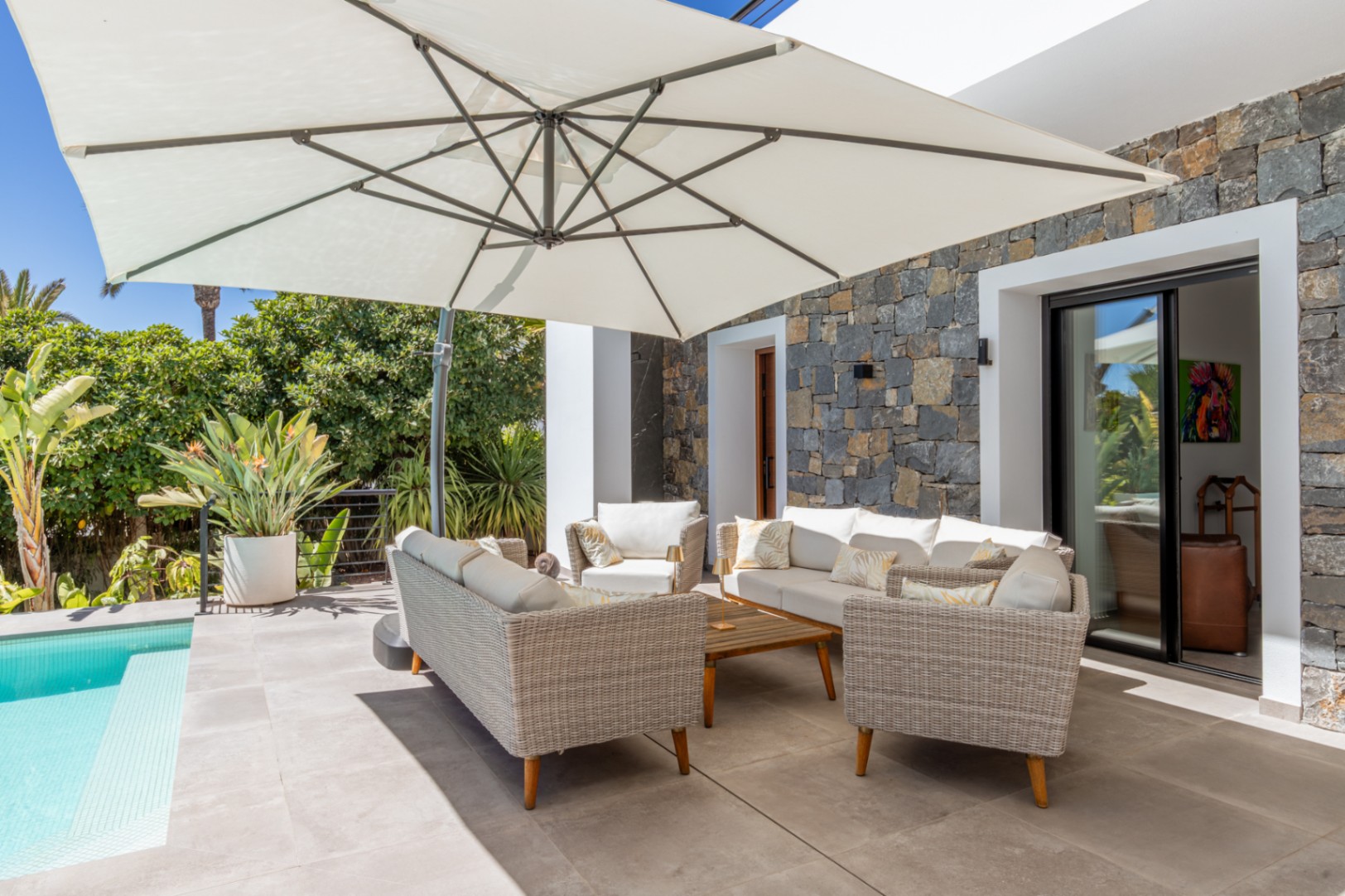 Outdoor lounge area featuring wicker furniture and a large cantilever umbrella, situated on a tiled terrace beside the pool with a natural stone facade.