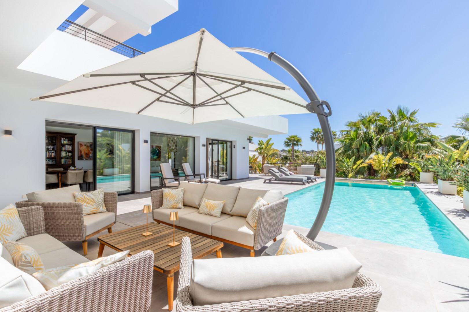 Outdoor lounge area with wicker seating and a large parasol by the pool, featuring direct access to the interior through large sliding glass doors.