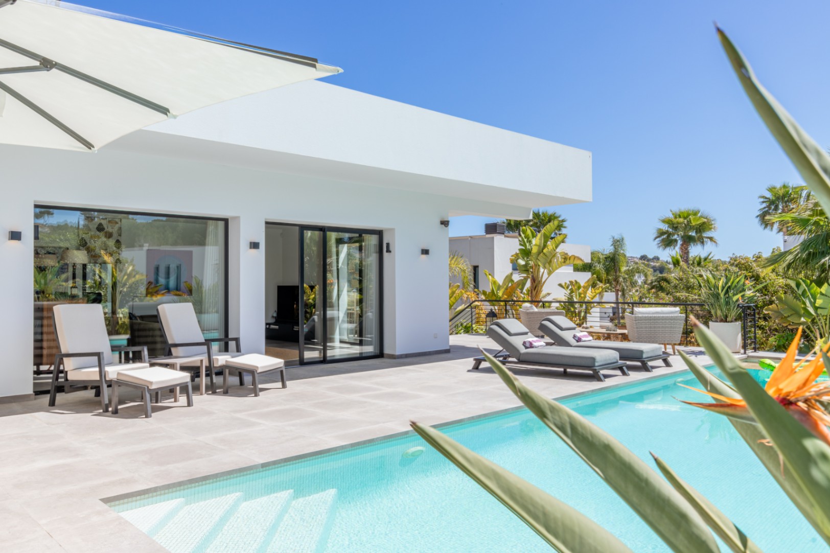 Outdoor terrace featuring a light-tiled pool, designer sun loungers, and direct living room access through black-framed sliding doors set against minimalist white architecture.