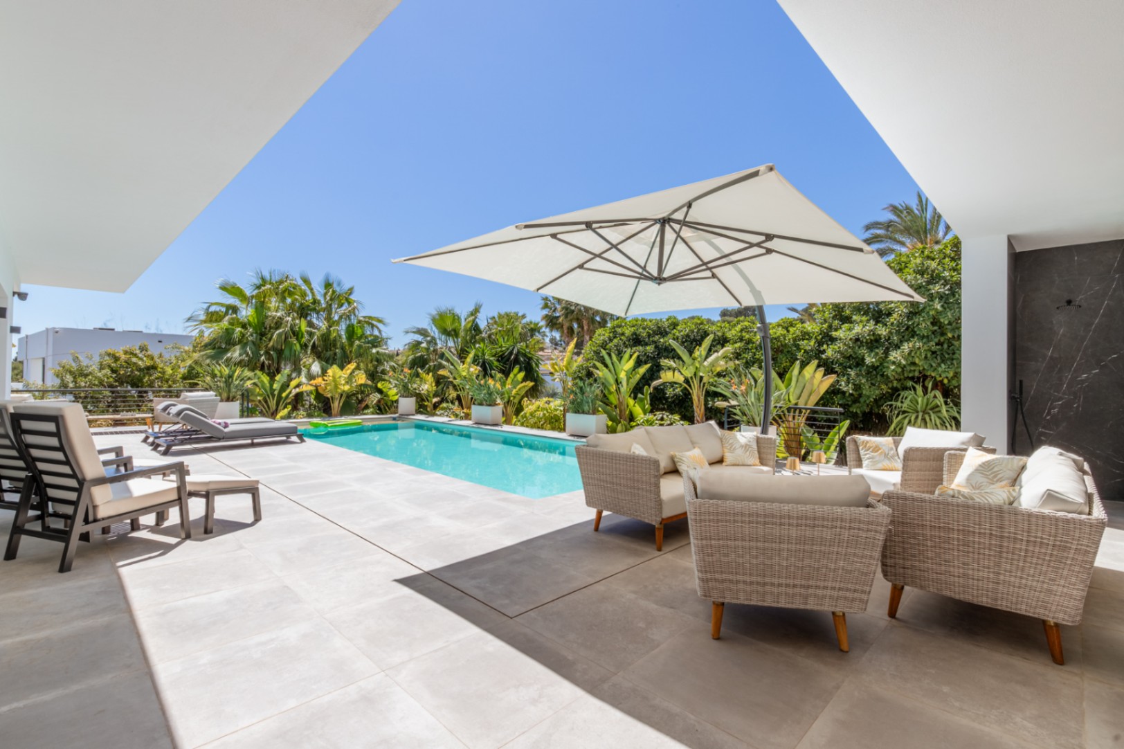 Outdoor terrace featuring a rectangular swimming pool, wicker lounge furniture under a cantilever umbrella, and lush Mediterranean landscaping for privacy.