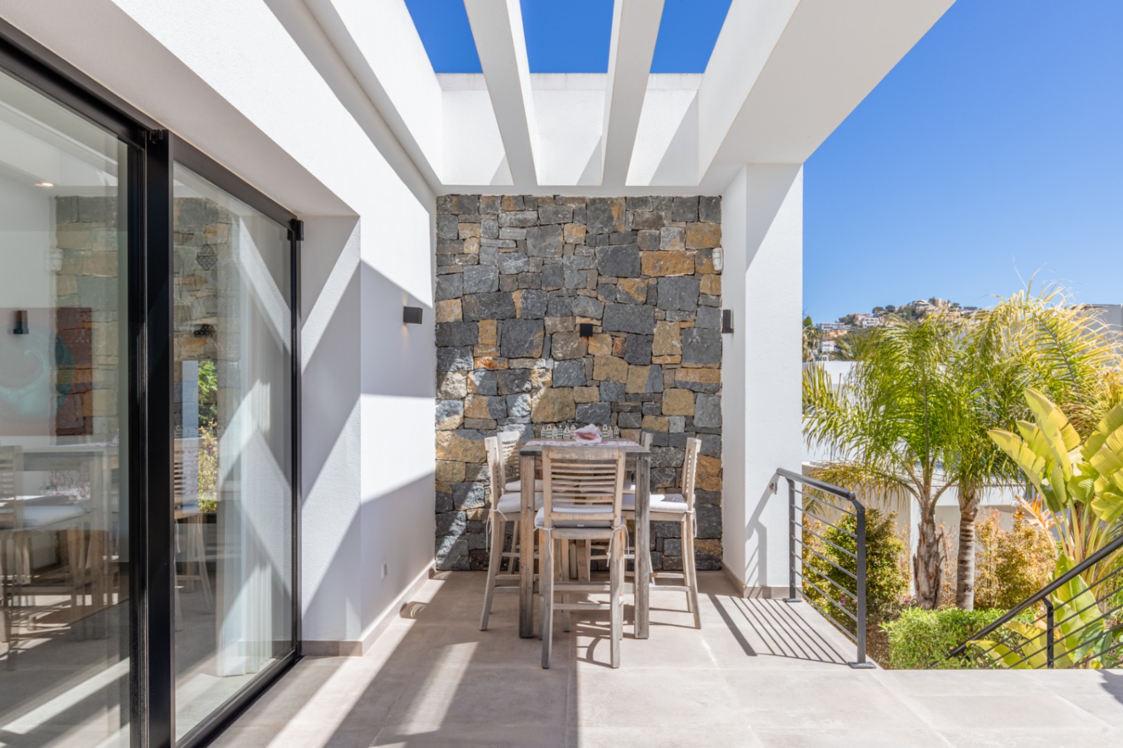 Outdoor terrace featuring a white pergola, natural stone accent wall, and high wooden dining set. Large glass doors connect the space to the interior under a clear sky.