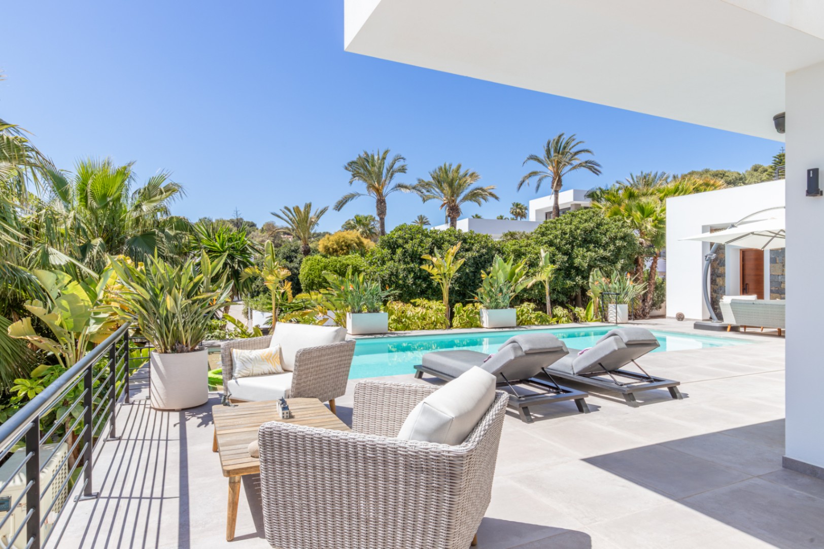 Outdoor terrace featuring wicker seating and sun loungers by the pool, set within a mature Mediterranean garden with palms and lush greenery.