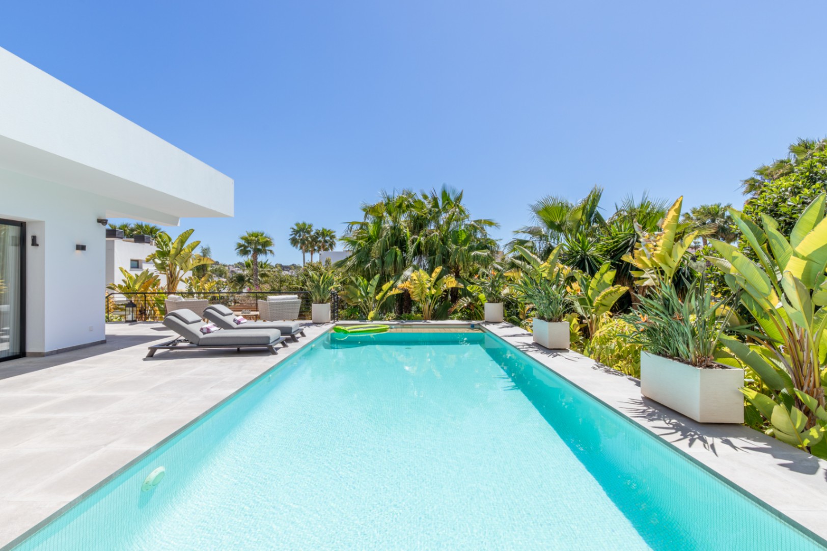 Rectangular swimming pool featuring grey porcelain tiling, bordered by mature tropical plants and modern sun loungers in a private, south-facing setting.