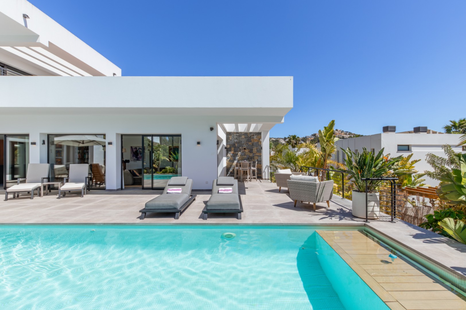 Spacious terrace featuring a turquoise tiled pool, sun loungers, and an outdoor dining area under a pergola, framed by modern white architecture and Mediterranean plants.