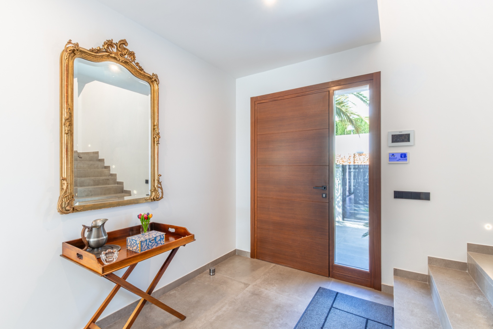 The entrance hall features a large wooden front door with a glass side panel, an ornate gold-framed mirror, and grey stone-effect floor tiles.
