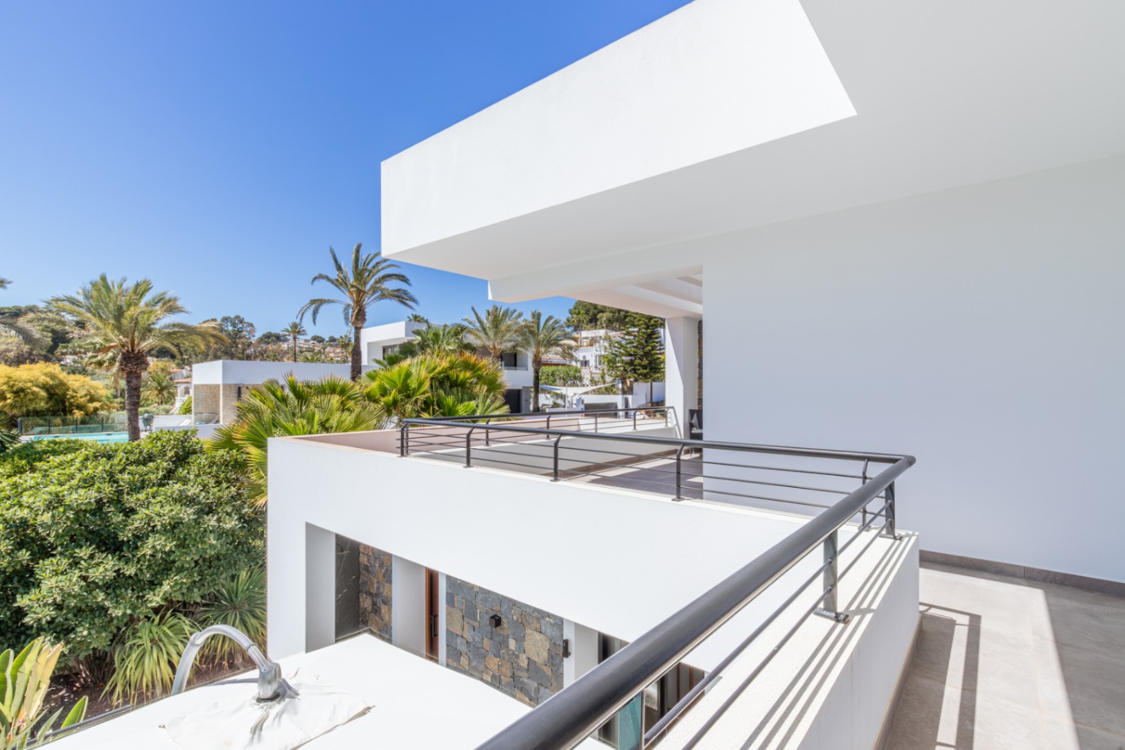 Upper-level terrace featuring a black metal railing and views of the Mediterranean garden. The modern white architecture incorporates natural stone accents and clean lines.