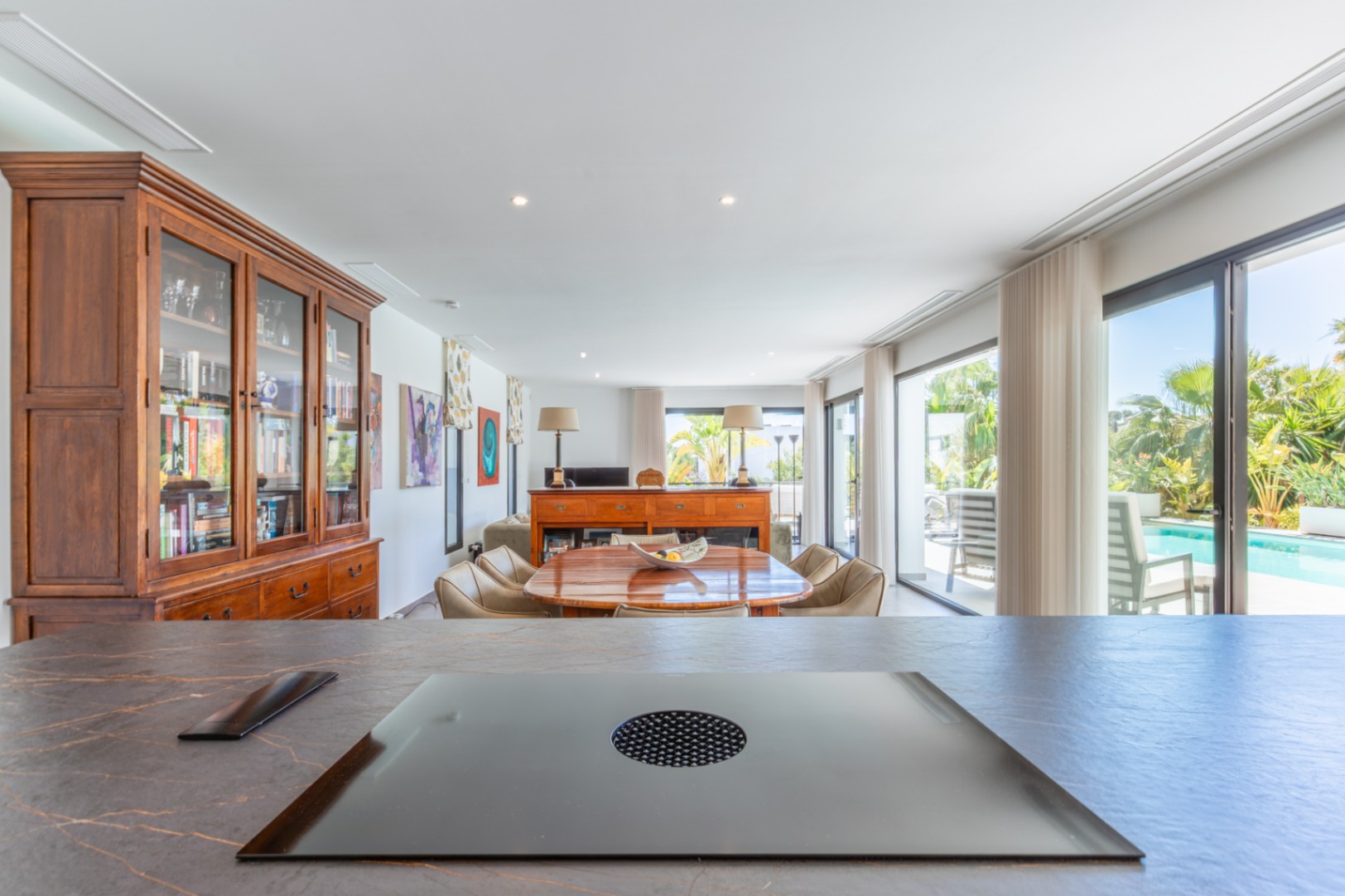 View from the kitchen island with integrated induction cooktop towards the dining area, featuring wooden furniture and floor-to-ceiling windows to the pool.