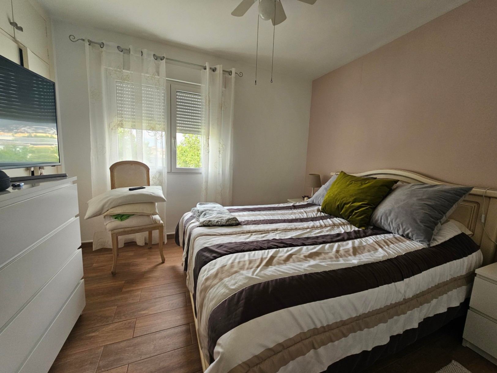 Bright bedroom featuring a pink accent wall, wood-effect tile flooring, a ceiling fan, and a window with integrated shutters and sheer curtains.