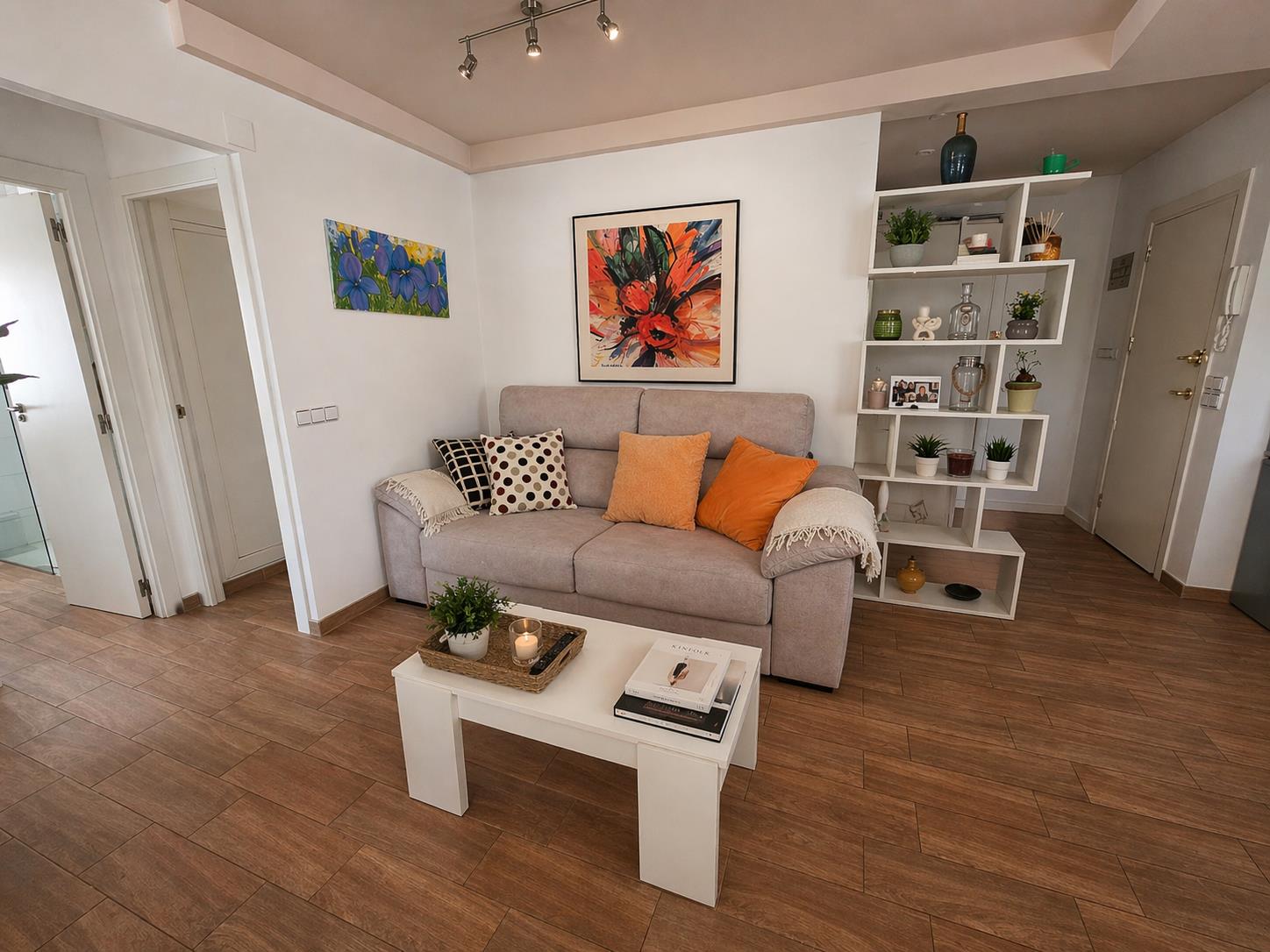 Cozy living area featuring a fabric sofa, white coffee table, and wood-effect tile flooring. A geometric shelving unit separates the space from the entrance.
