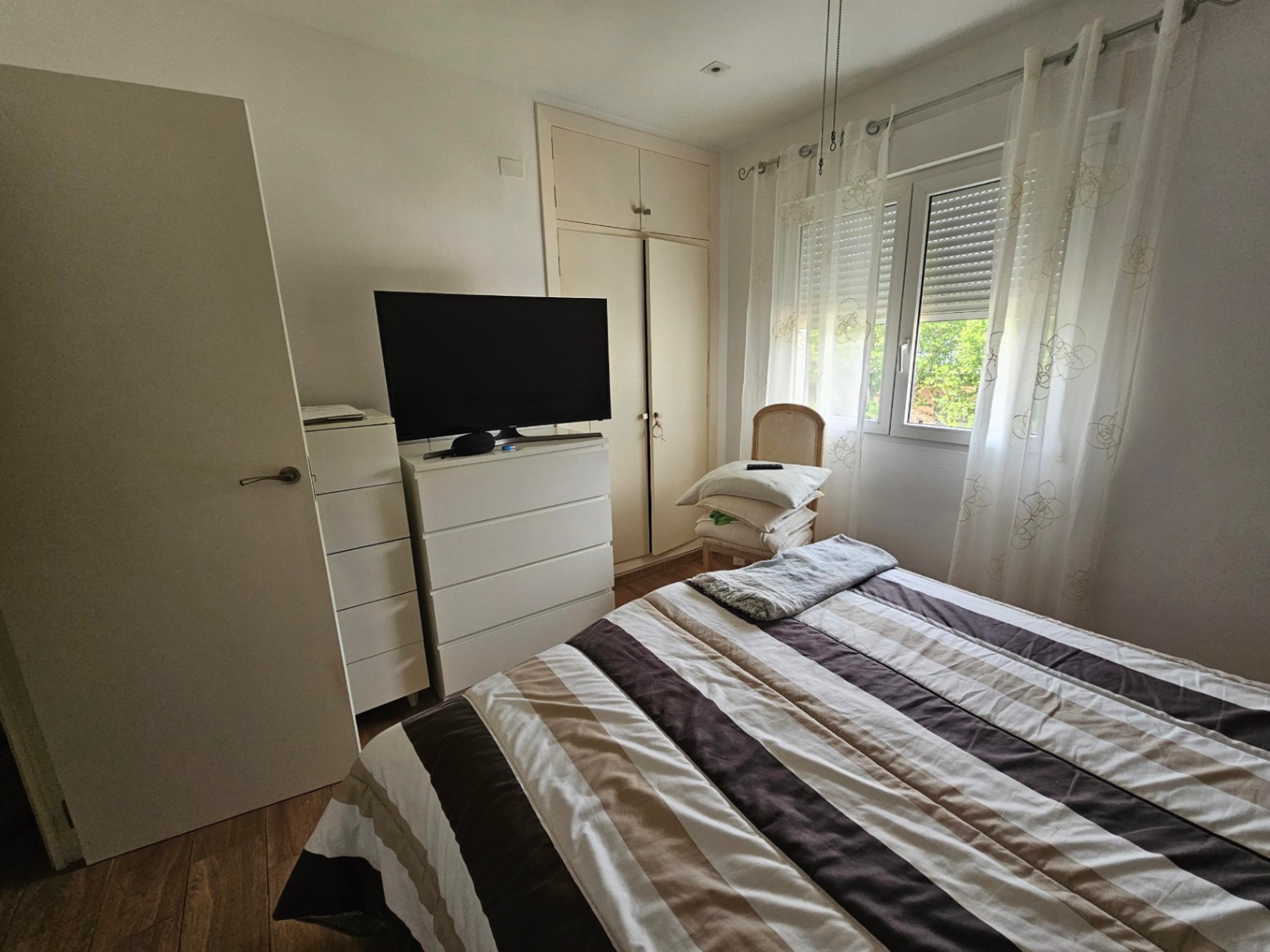 Functional bedroom featuring built-in wardrobes, white dressers, and a window with integrated shutters providing natural light over laminate flooring.