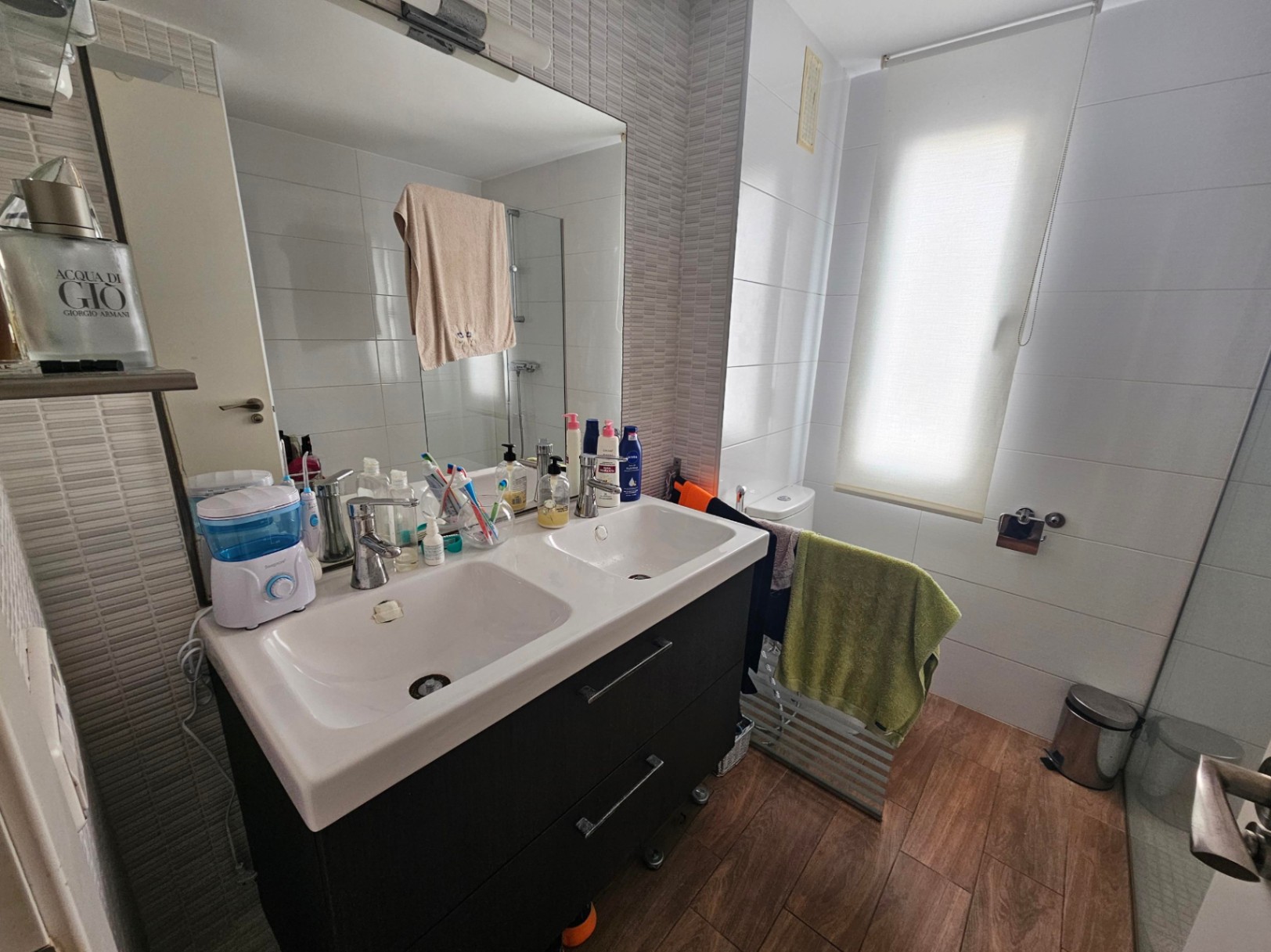 Modern bathroom featuring a double vanity with dark cabinetry, wood-effect porcelain flooring, and neutral wall tiling illuminated by natural light.