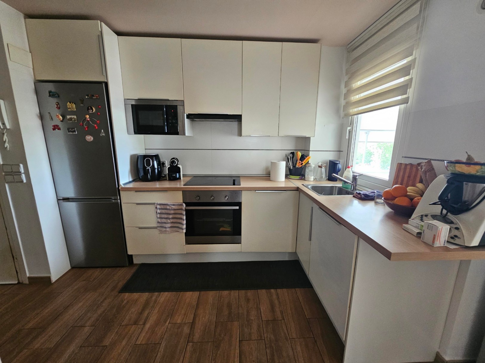 Modern L-shaped kitchen featuring white cabinetry, light wood-effect countertops, and stainless steel appliances complemented by wood-look tiled flooring.
