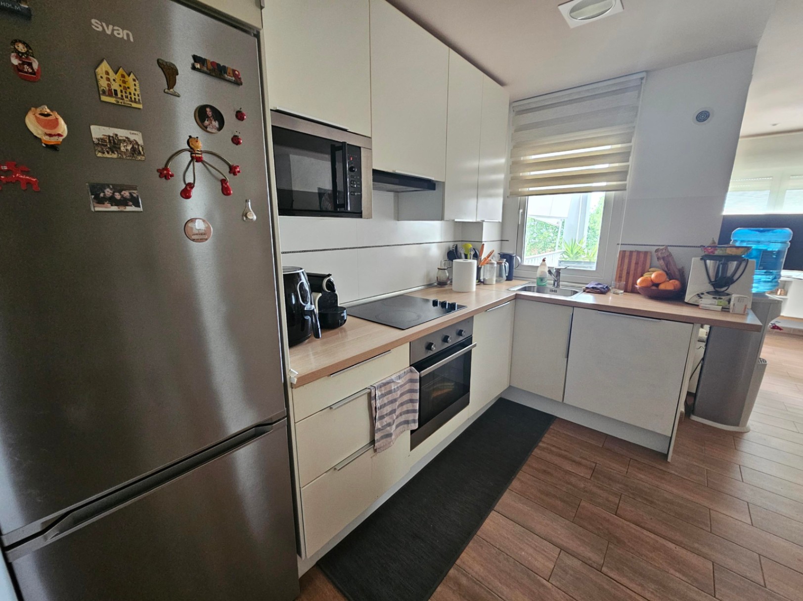 Modern L-shaped kitchen featuring wood-effect countertops, integrated stainless steel appliances, and natural light from a window with blinds.