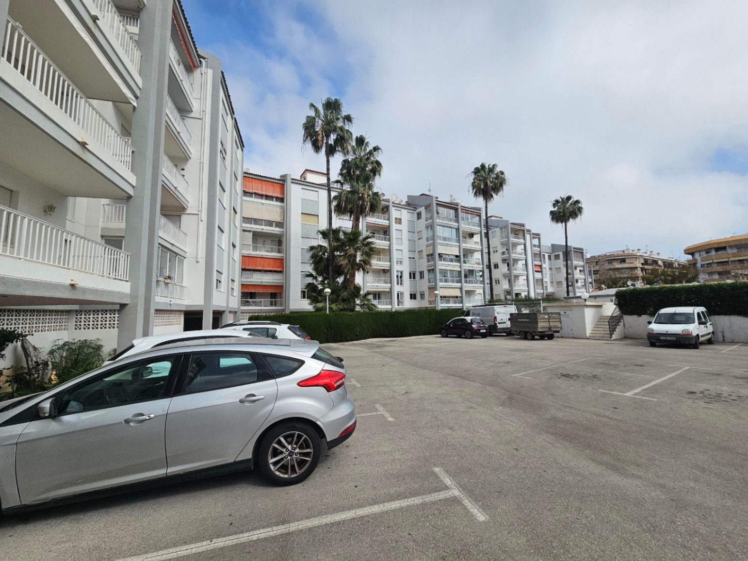 Outdoor communal parking area with marked spaces, surrounded by manicured hedges and palm trees in front of the residential building facade.