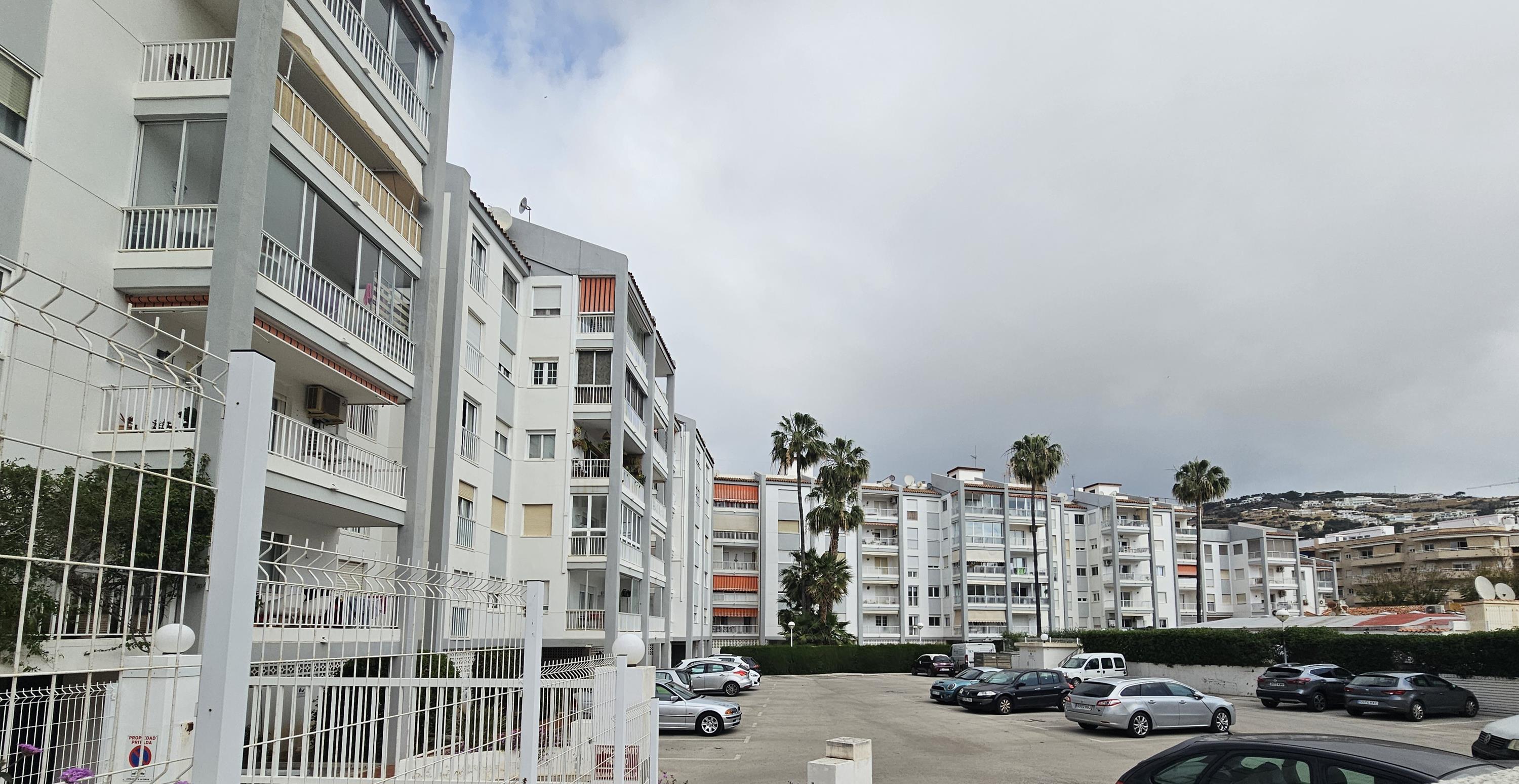 Residential building facade with balconies and awnings, featuring a large gated communal parking area and landscaped areas with palm trees.
