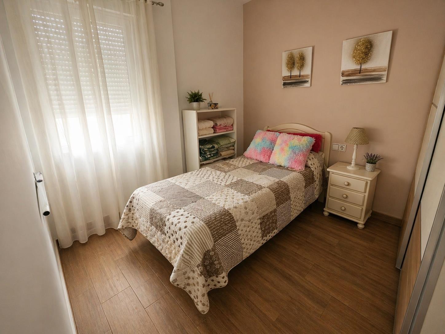 Single bedroom featuring wood-effect flooring, a window with shutters and white curtains, and a warm-toned accent wall.