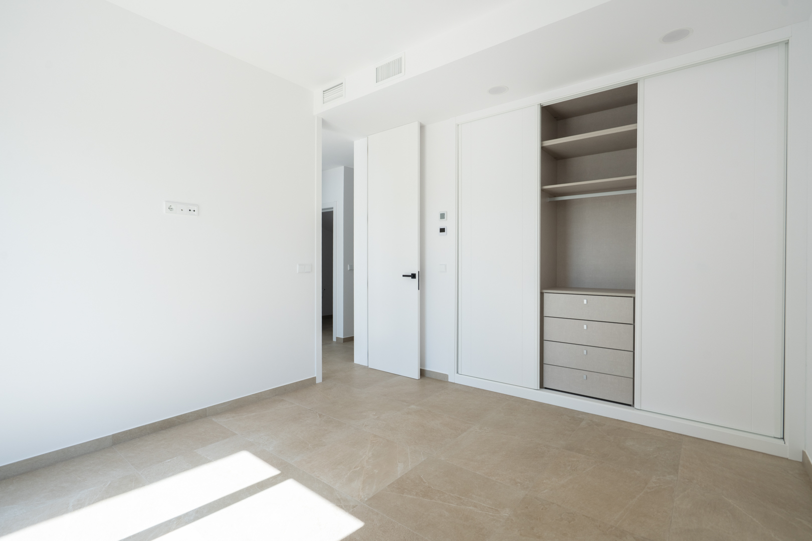 Bright bedroom featuring built-in wardrobes with white sliding doors, integrated shelving and drawers, and large-format porcelain stone-effect flooring.
