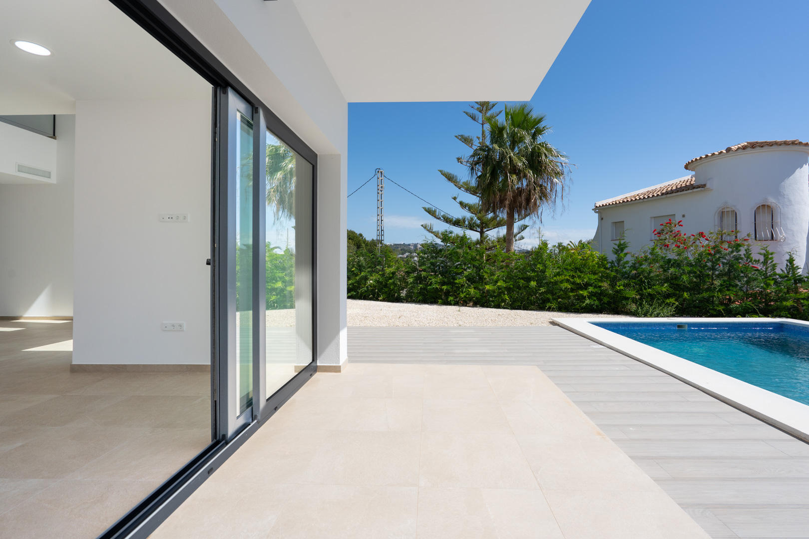 Covered terrace featuring porcelain flooring and large sliding glass doors that open to a swimming pool area with wood-effect decking and Mediterranean greenery.
