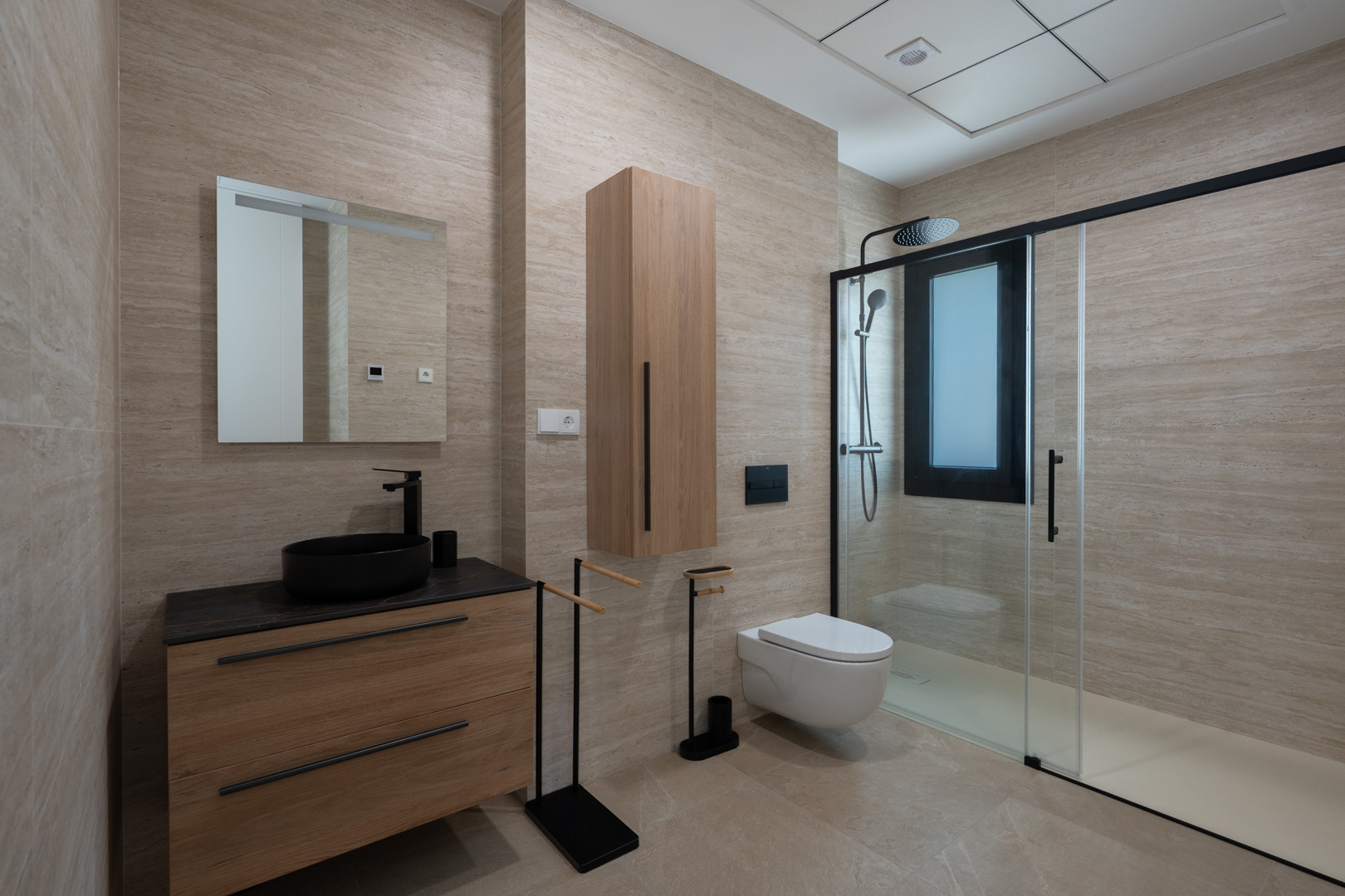 Designer bathroom featuring travertine-effect porcelain tiling, a wooden vanity with a black vessel sink, and a walk-in shower with sleek black framing.