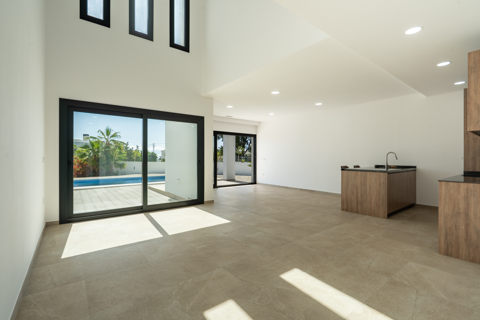 Double-height open-plan living area featuring a designer kitchen island, porcelain flooring, and large sliding doors opening to the pool terrace.