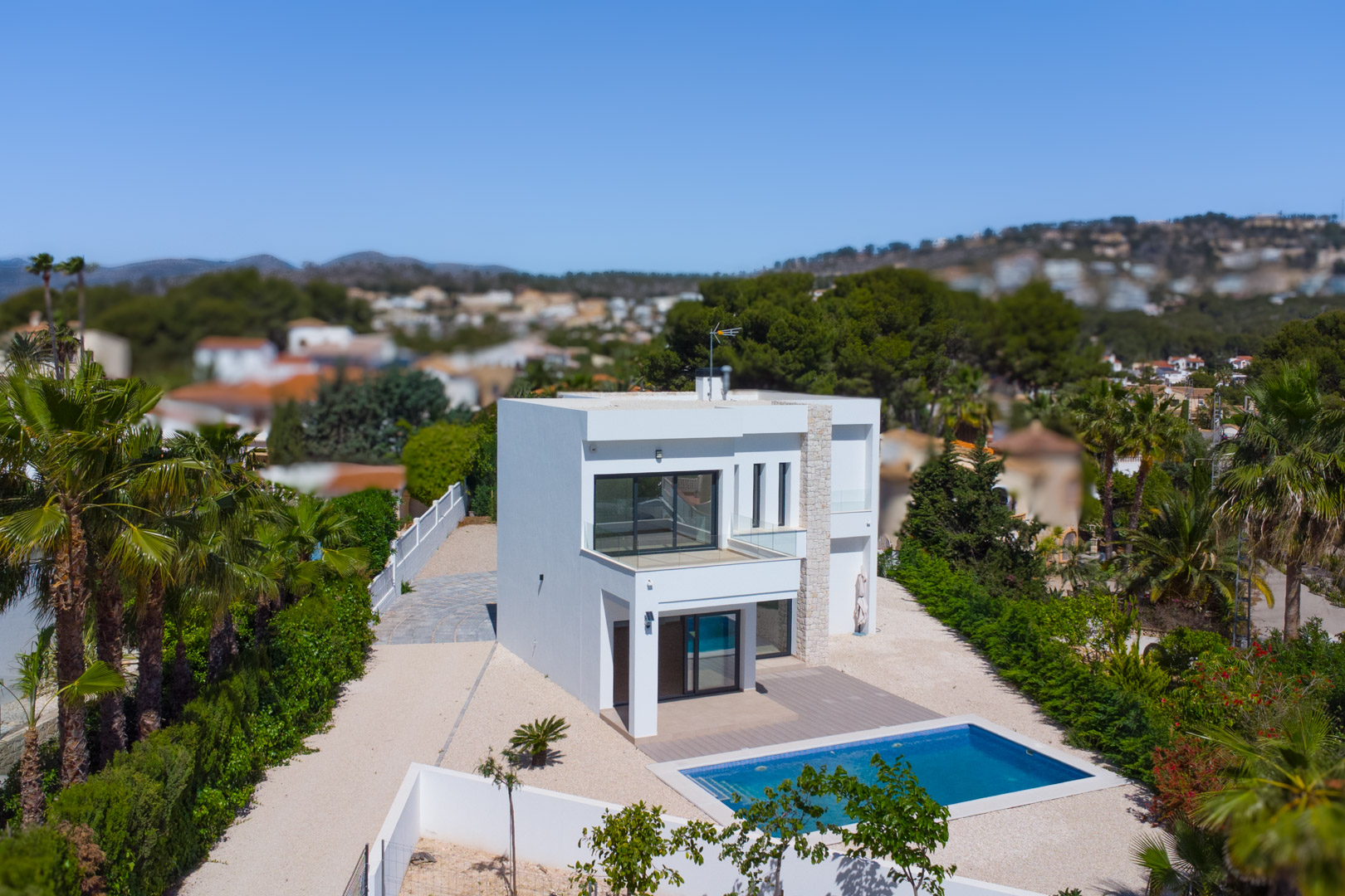 Exterior view of the modern villa featuring a white facade, natural stone accents, a rectangular pool, and glass-railed terraces on an expansive flat plot.