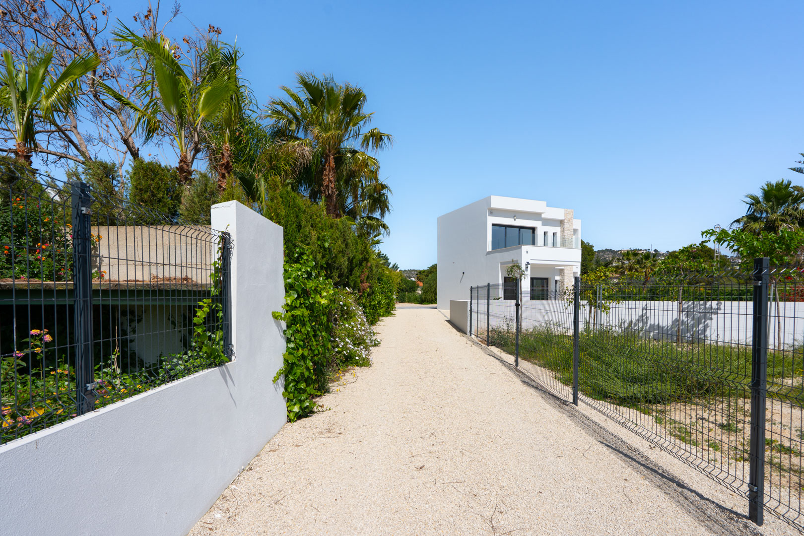 Long gravel driveway leading to the modern white villa, featuring perimeter fencing, Mediterranean palms, and an expansive flat plot area.
