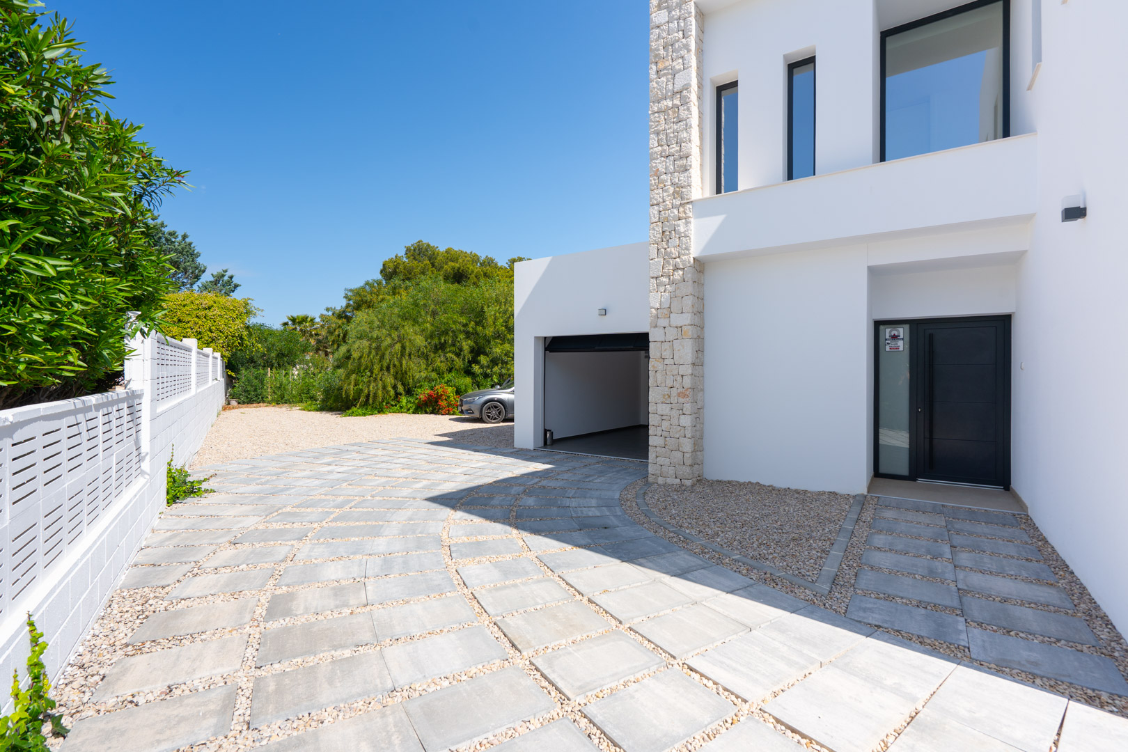 Main entrance and garage access featuring a tiled driveway with gravel accents, complemented by a white facade and a natural stone pillar.