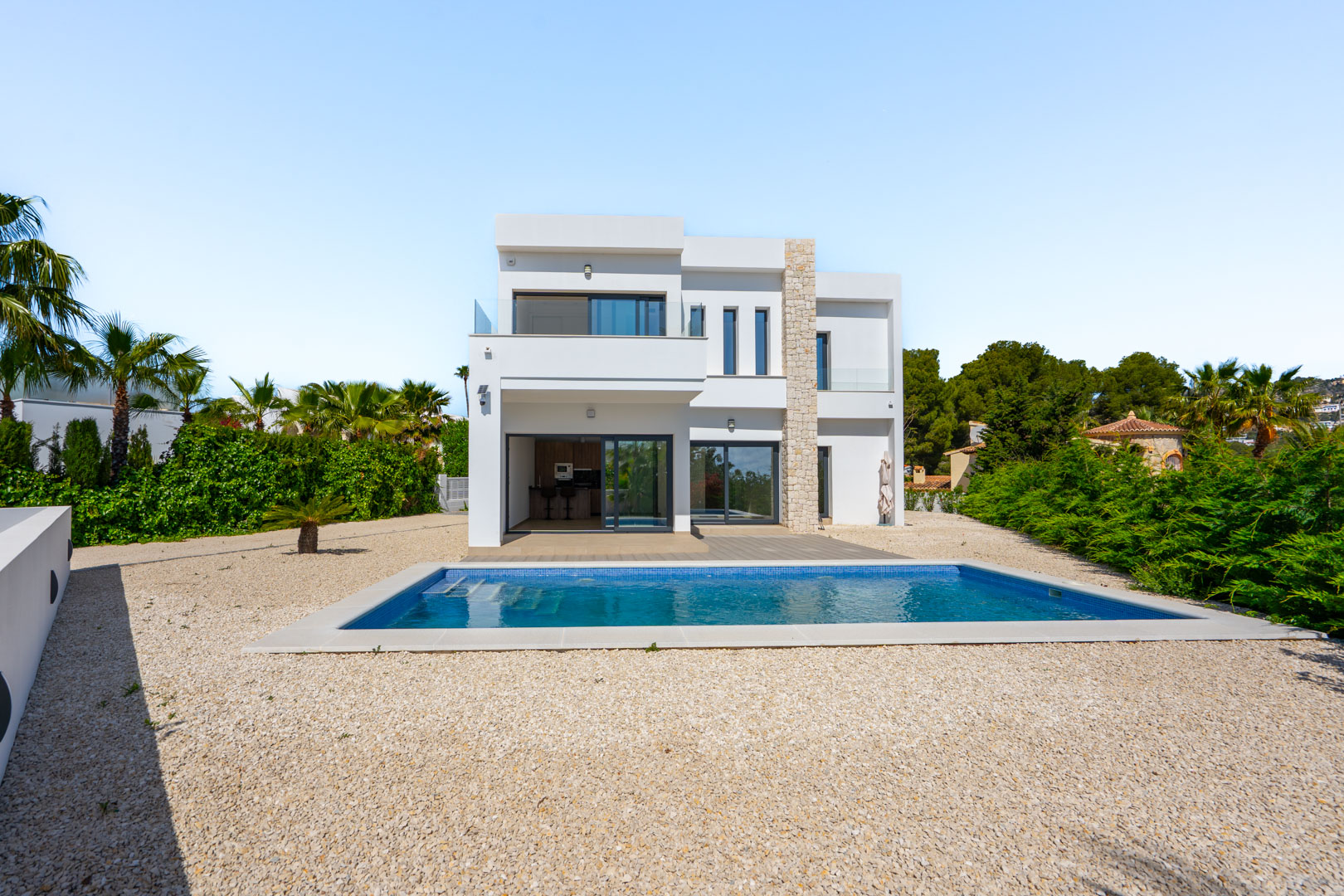 Modern two-story villa featuring a white facade with natural stone accents, a rectangular swimming pool, and gravel landscaping under a clear sky.