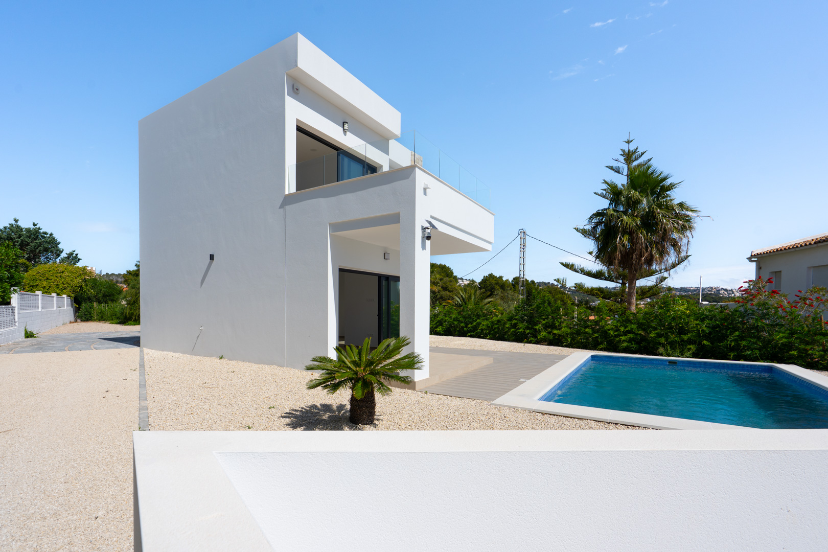 Modern white villa exterior featuring a rectangular swimming pool, minimalist gravel landscaping, and upper-level terraces with glass balustrades.